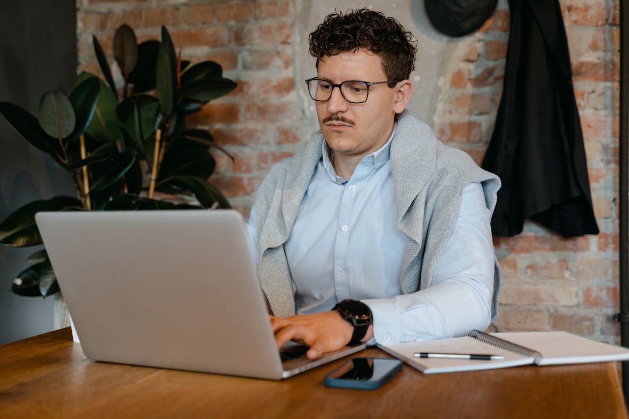 A young man wearing glasses works on a laptop in a modern office setting.